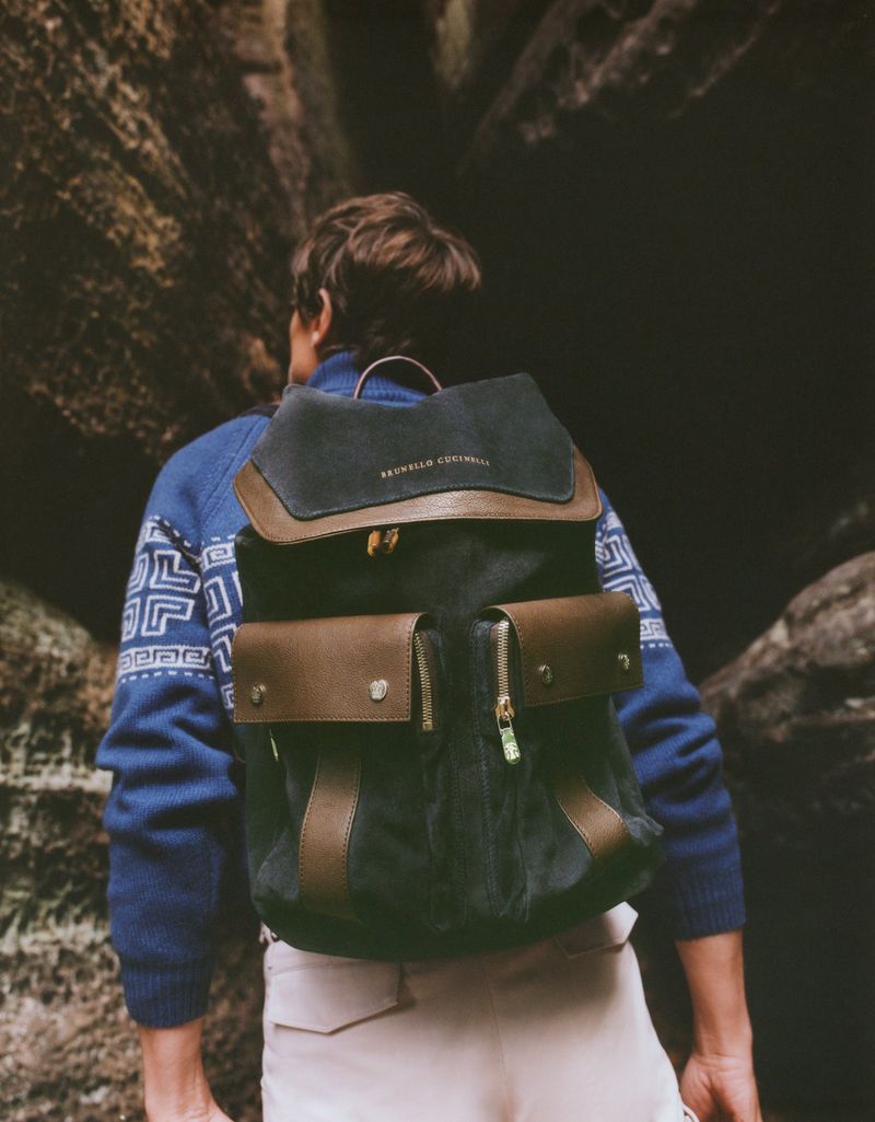 A man wearing a blue patterned sweater and carrying a dark suede backpack standing between rocky canyon walls.