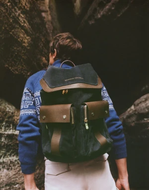 A man wearing a blue patterned sweater and carrying a dark suede backpack standing between rocky canyon walls.