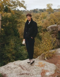 Model standing on a rocky outcrop wearing a dark peacoat and dark trousers against a dense autumn woodland landscape.