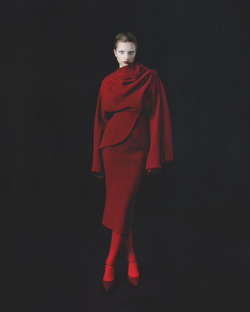 Model Kristin Drab poses in a structured crimson wrap jacket, midi skirt, red tights, and matching heels against a black studio background.