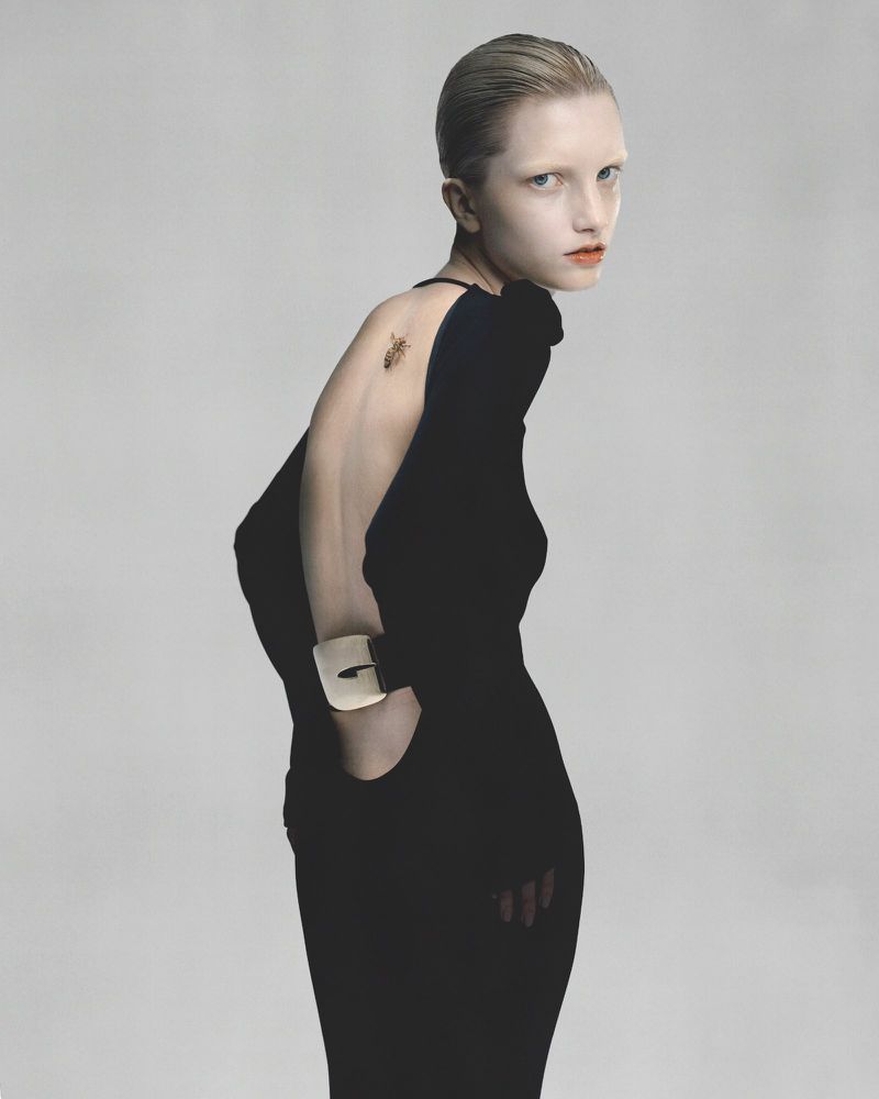 A woman in a dark dress with an open back glances over her shoulder in a studio fashion shot, highlighted by clean neutral background.