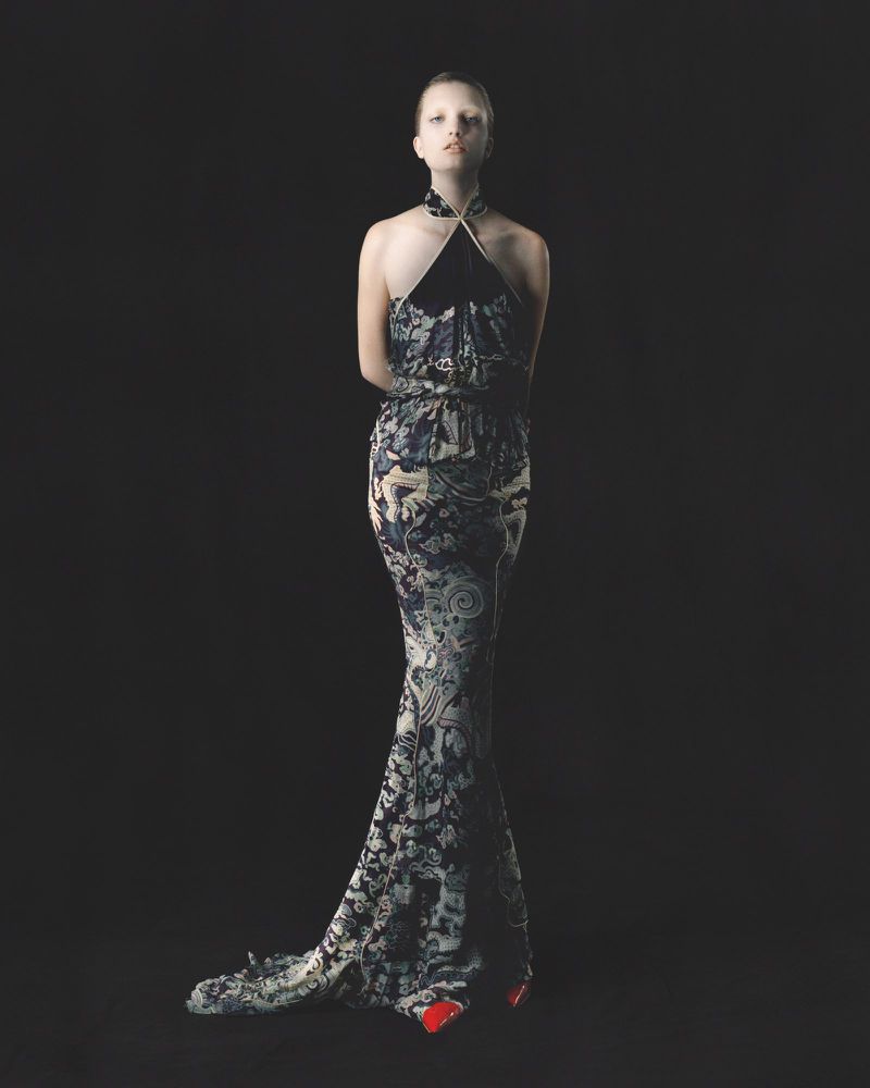 Fashionable model wearing a full-length halter gown with a floral pattern, posing against a dark backdrop in a studio setting.