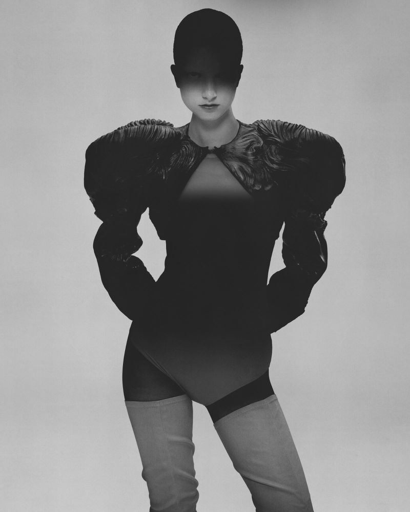 Woman poses in a bodysuit with oversized sleeves, matched with thigh-high boots. The backdrop is light grey, emphasizing the dark outfit.