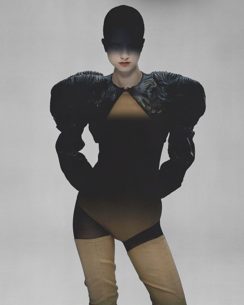 A woman in an avant-garde black bodysuit with puffy shoulders poses against a plain gray studio backdrop in a fashion photography setting.