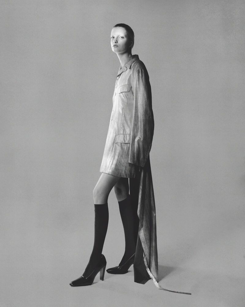 Monochromatic studio shot of a slim, fair-skinned adult posing in a striped dress, black knee-high socks, and high heels, against a gray background.