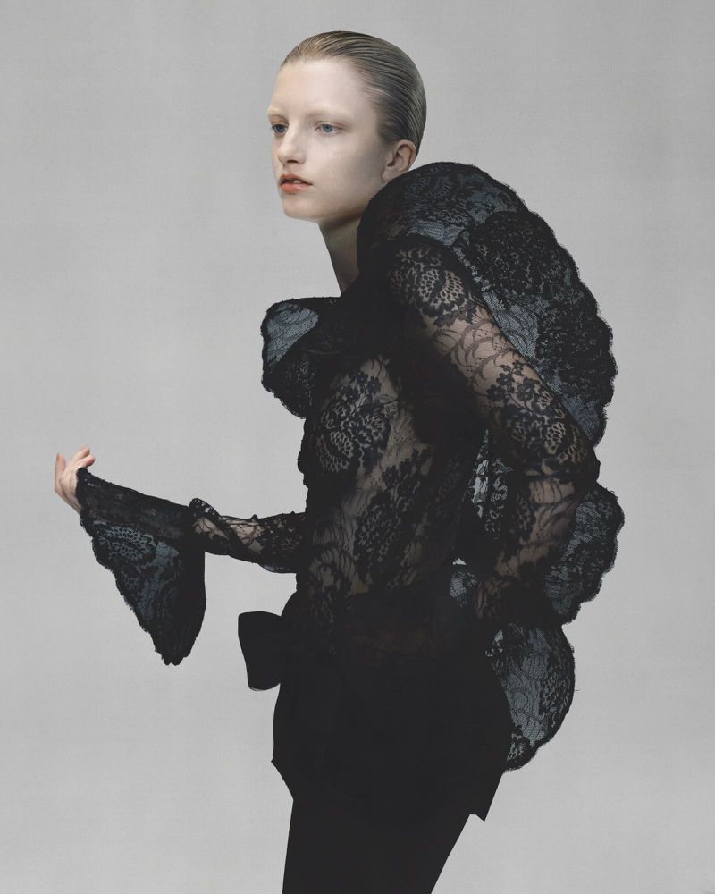 Fashion shot of a woman with slicked-back hair wearing a black lace top with exaggerated shoulders and standing against a grey background.