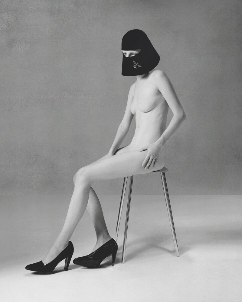 Model wearing a black sculptural headpiece and black high heels seated on a minimalist stool in a studio setting.