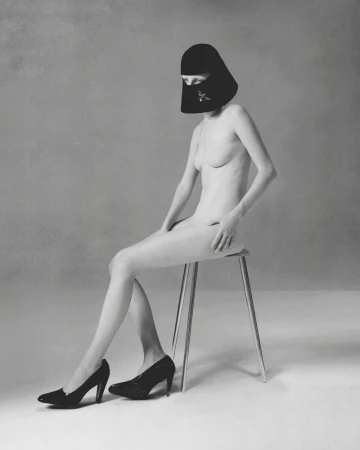 Model wearing a black sculptural headpiece and black high heels seated on a minimalist stool in a studio setting.