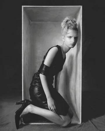 Model Kristin Drab kneeling inside a rectangular open box wearing a black dress and heels in a black and white editorial portrait.