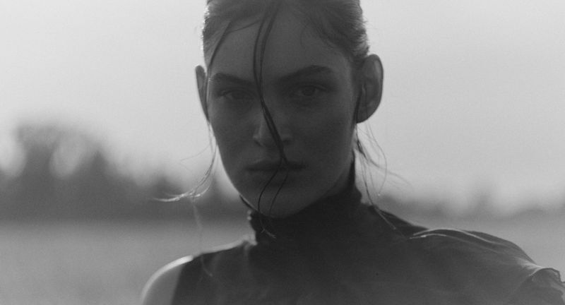 Monochrome portrait of a woman with wet hair, looking directly at the camera against a blurred outdoor backdrop with neutral tones.