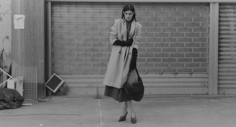 A woman in a coat stands in front of a brick wall and metal garage door with a handbag on her shoulder in a black and white fashion shot.