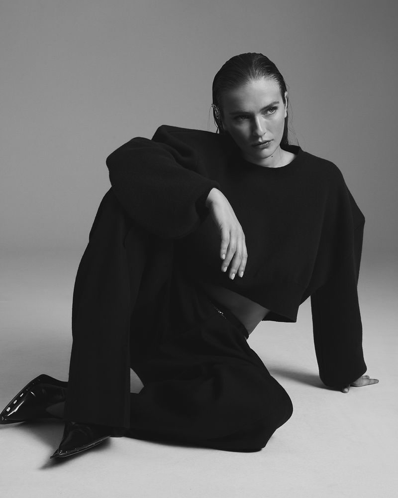 Black and white shot of a female model in a relaxed pose, wearing a black cropped sweater, wide-leg pants, and patent leather shoes.