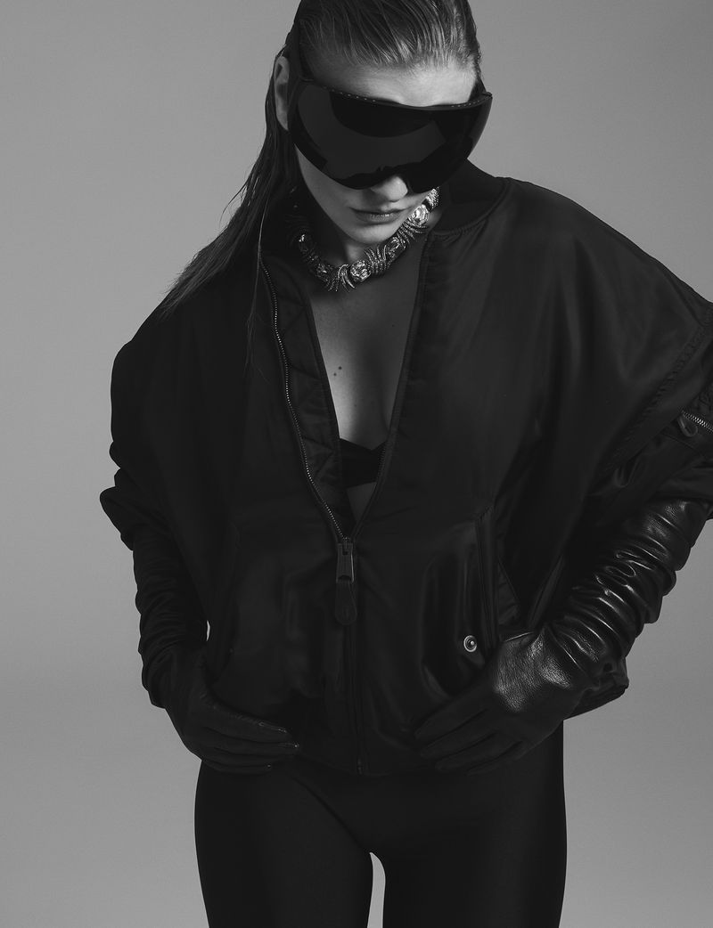 Camille Razat wearing oversized visor sunglasses, an unzipped black jacket, and long black leather gloves against a grey backdrop.