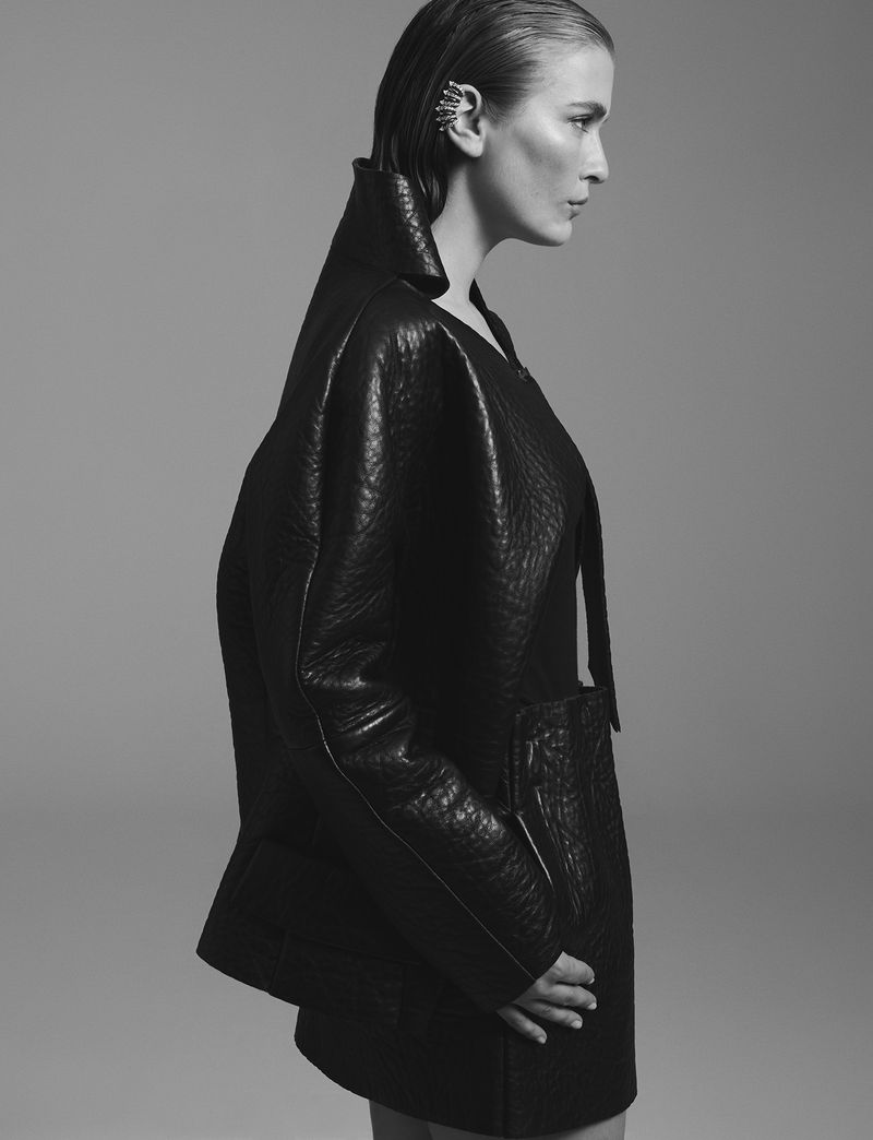 Profile shot of an adult woman in a black leather jacket and wet hair, wearing an ear cuff, against a solid light grey background.
