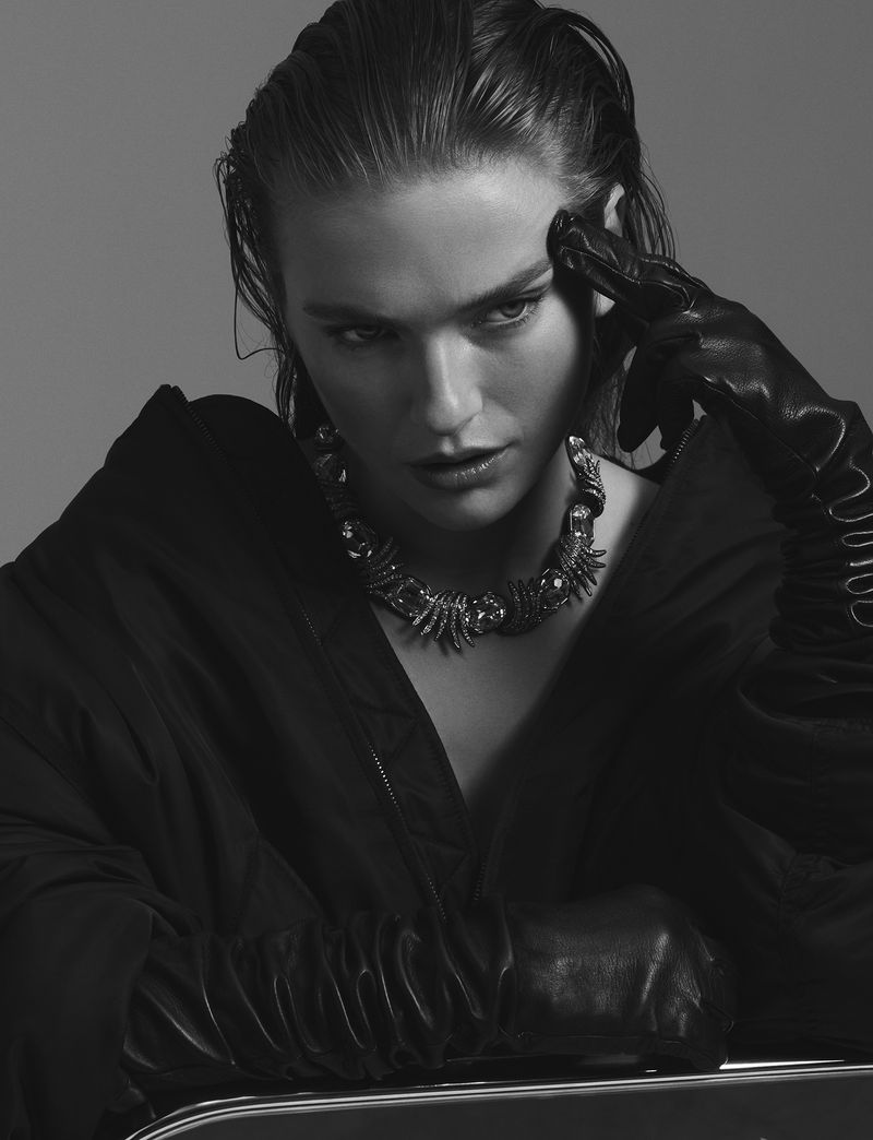 Monochromatic portrait of an adult female model with wet hair, wearing a sculptural necklace, leather gloves, and a black jacket.