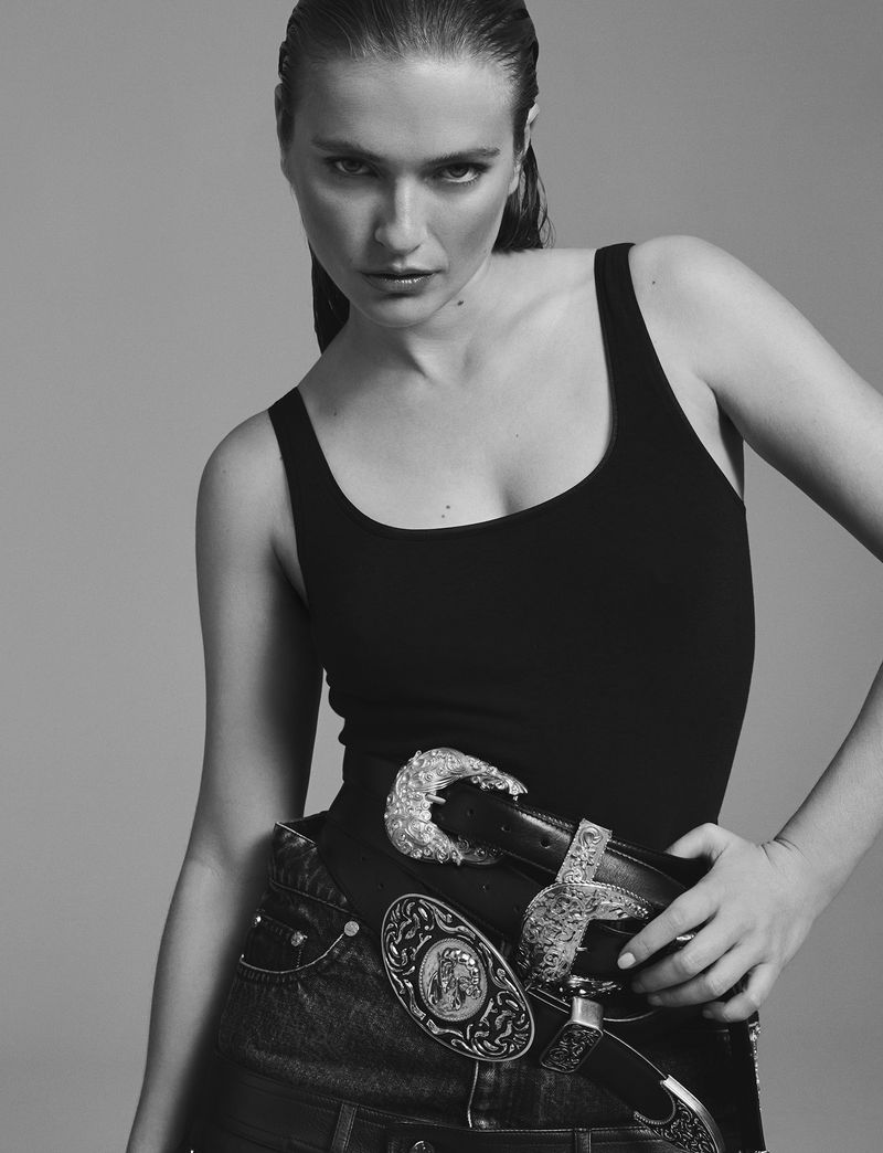 Black and white fashion portrait of a woman in a black tank top, jeans, and decorative belts looking at the camera in a studio.