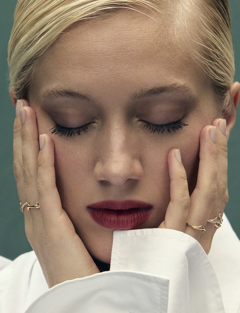 Close-up shot of a fair-skinned woman with blonde hair, eyes closed, wearing a white shirt and a gold ring, set against a teal backdrop.