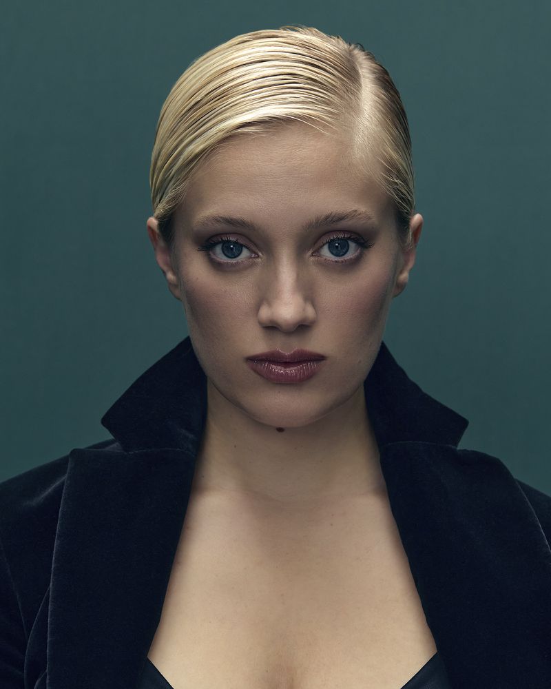 Close-up portrait of Nadia Tereszkiewicz with sleek blonde hair in a black velvet jacket against a teal background.