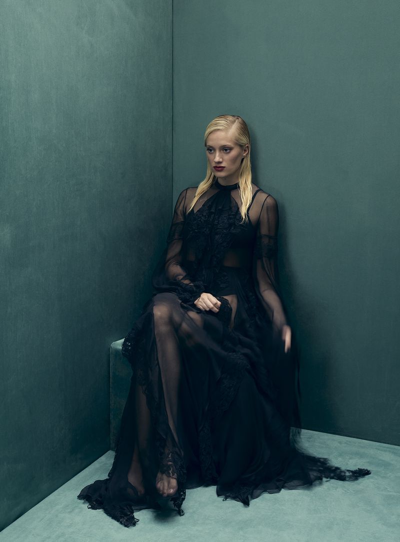 Elegant woman in a dark, sheer gown sits gracefully in a corner with teal walls, illuminated by soft, diffused lighting in a fashion editorial.