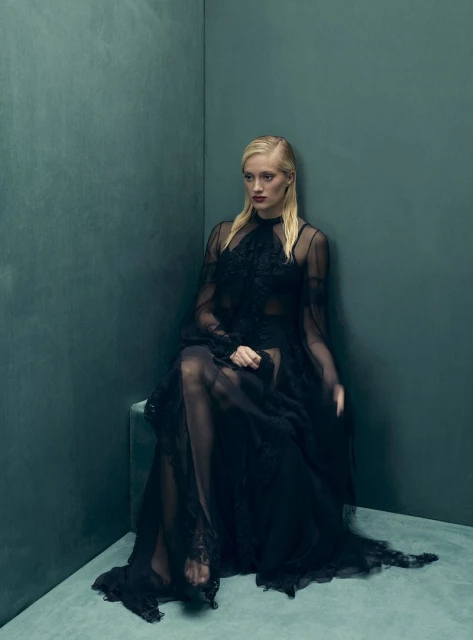 Elegant woman in a dark, sheer gown sits gracefully in a corner with teal walls, illuminated by soft, diffused lighting in a fashion editorial.