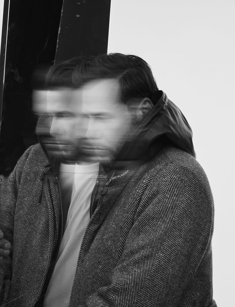 Monochrome portrait of a man wearing a herringbone coat with a white shirt, captured with a long-exposure technique, showing motion blur.