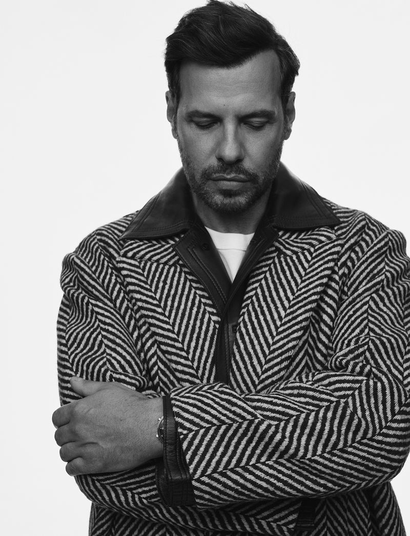Monochrome portrait of a man with dark hair and a patterned coat, standing indoors against a neutral background, eyes downcast.