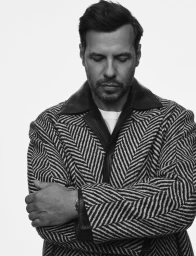 Black and white portrait of actor Laurent Lafitte looking downward with arms crossed, wearing a chevron coat with leather collar.