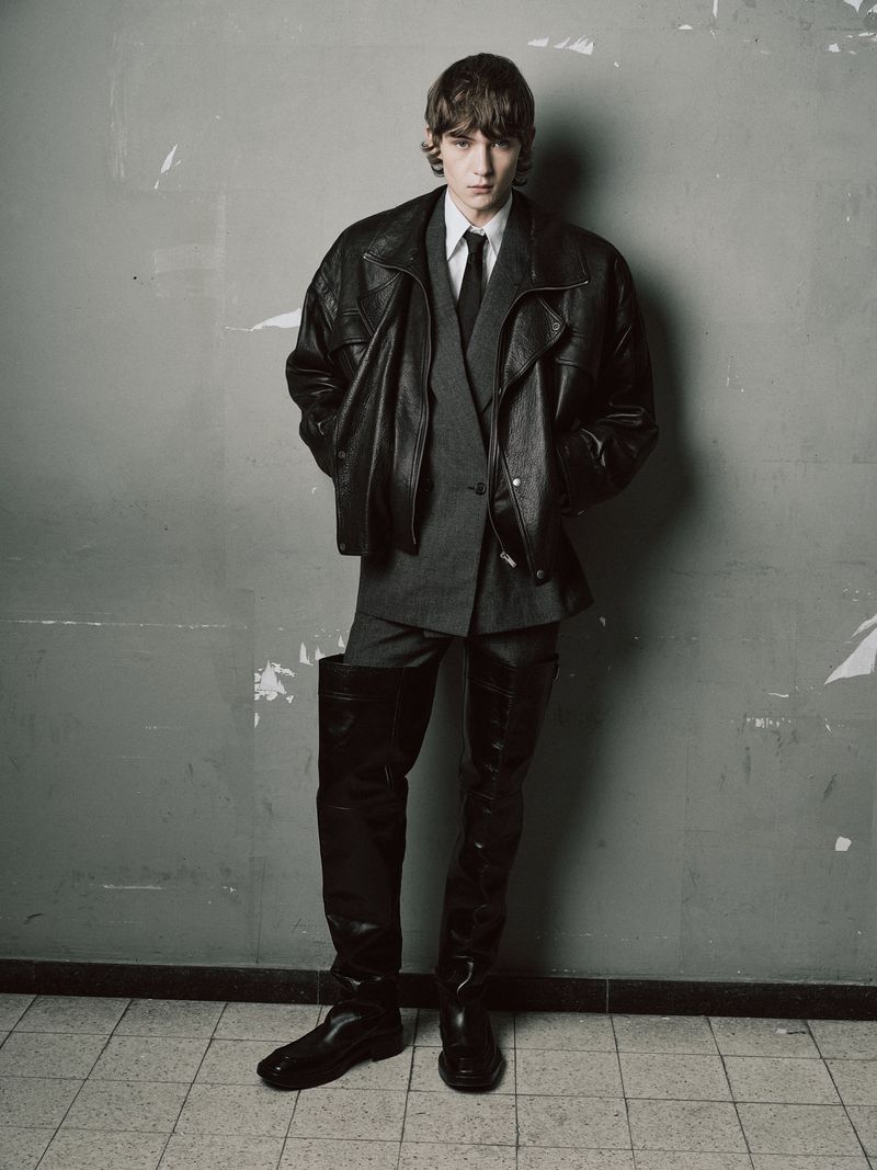 A stylish adult male poses in a black leather jacket, grey suit, black tie, and black boots against a textured grey wall and tiled floor.