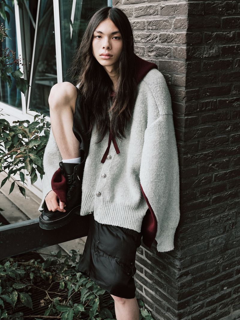 Model with long dark hair wearing a grey oversized cardigan and black boots, leaning against a brick pillar.