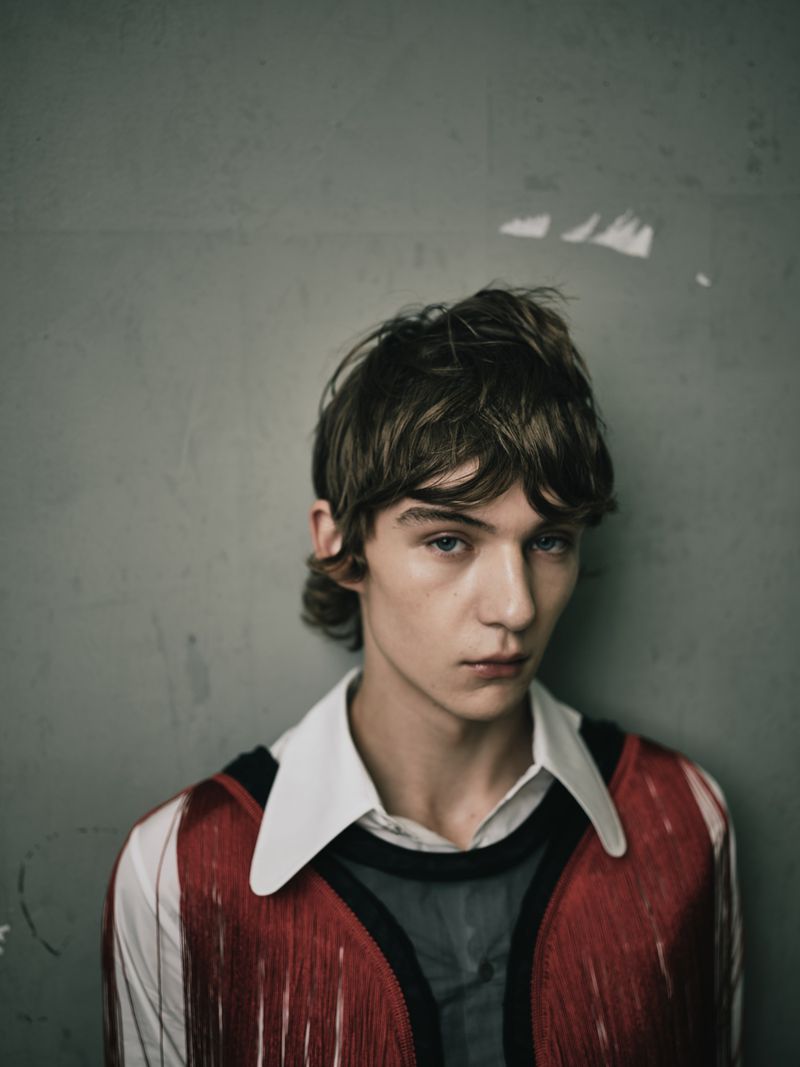 Portrait of a young adult man with brown hair and blue eyes, wearing a white collared shirt and fringed red vest against a grey background.