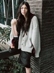 Model with long dark hair wearing a grey oversized cardigan and black boots, leaning against a brick pillar.