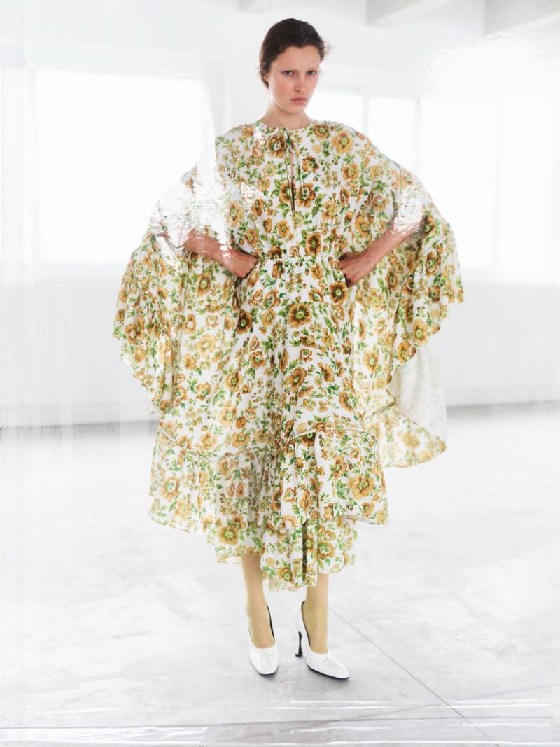 A slender woman poses in a white studio wearing a vintage-style floral dress and white heels, standing in front of large windows.