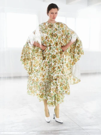 A slender woman poses in a white studio wearing a vintage-style floral dress and white heels, standing in front of large windows.