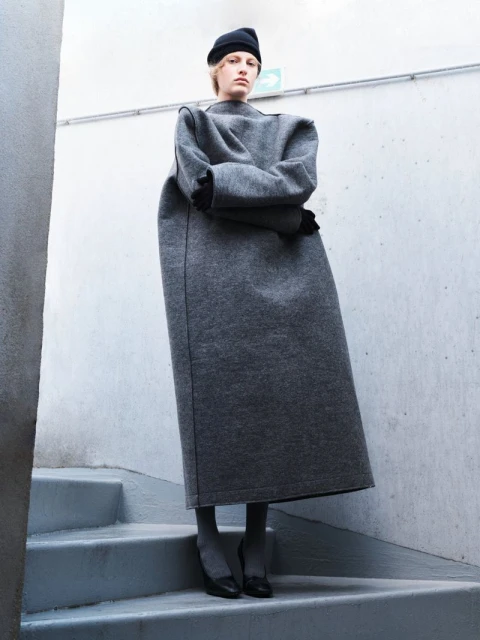 Fashion shot of a woman wearing a grey, oversized dress and a black hat, posing on grey stairs against a neutral wall indoors.