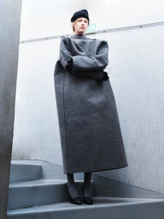 Model Felice Nova Noordhoff posing on concrete stairs in an oversized grey sculptural maxi coat and black beanie.