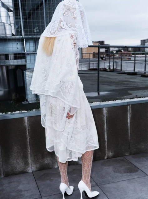 A woman in a white lace dress, veil, and high heels stands on a rooftop with a cloudy sky in the background, showcasing fashion and style.