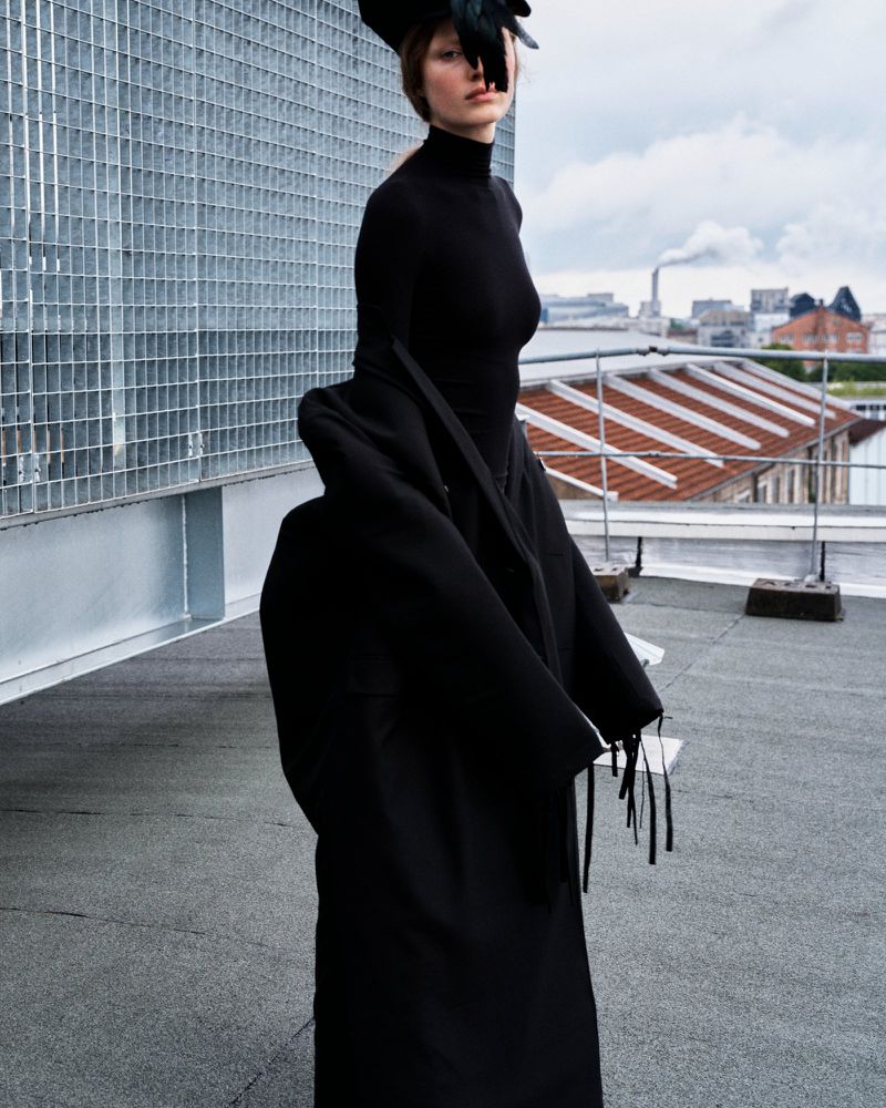 A model poses on a rooftop wearing a black dress and feathered headpiece, a dark coat, and with an urban skyline in the background.