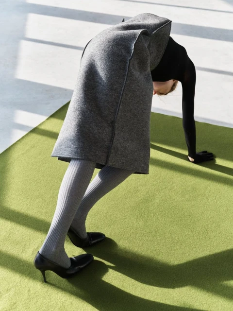 Adult woman in black top, grey skirt, tights, heels is doing a backbend on a green rug inside a studio with light coming through the window.