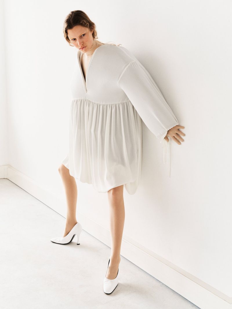 Woman in a loose white dress and white heels posing against a bright white wall. Fashion shot on bright neutral background.