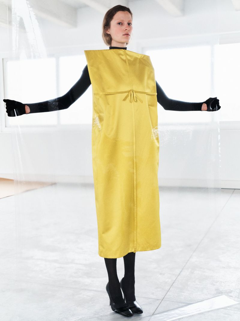 Fashion model in avant-garde yellow dress with black gloves and leggings, posing in minimalist white studio, creating a high fashion editorial.