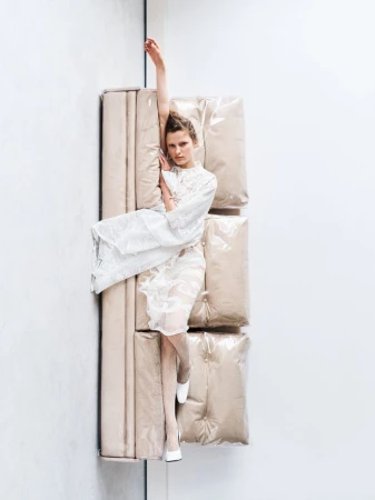 Fashion shot of a young adult woman in a sheer white dress, lying on a stack of three beige, cushion-like objects against a plain white wall.