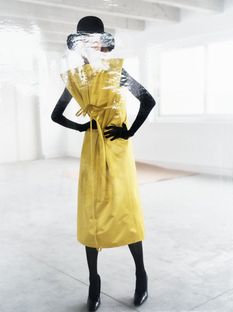 A stylish woman in a yellow dress, black gloves, and black hat poses in a minimalist white room with a blurred, dreamlike quality.