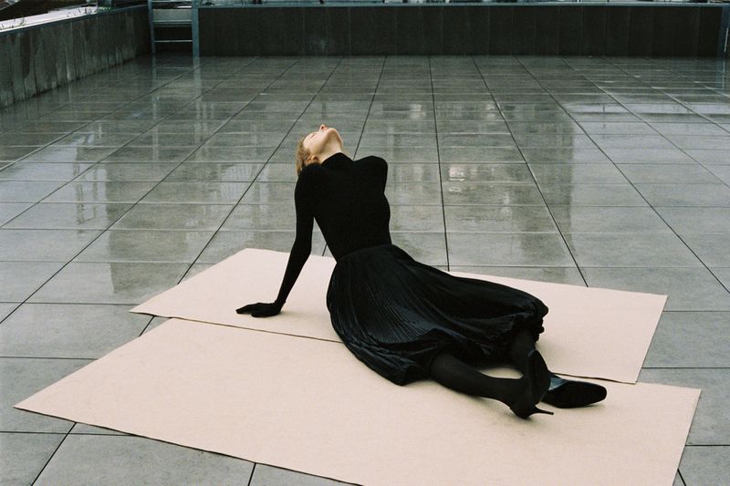 Woman in black lying on cardboard in an urban environment. She is reclining looking upwards with her head tilted back on grey surface.
