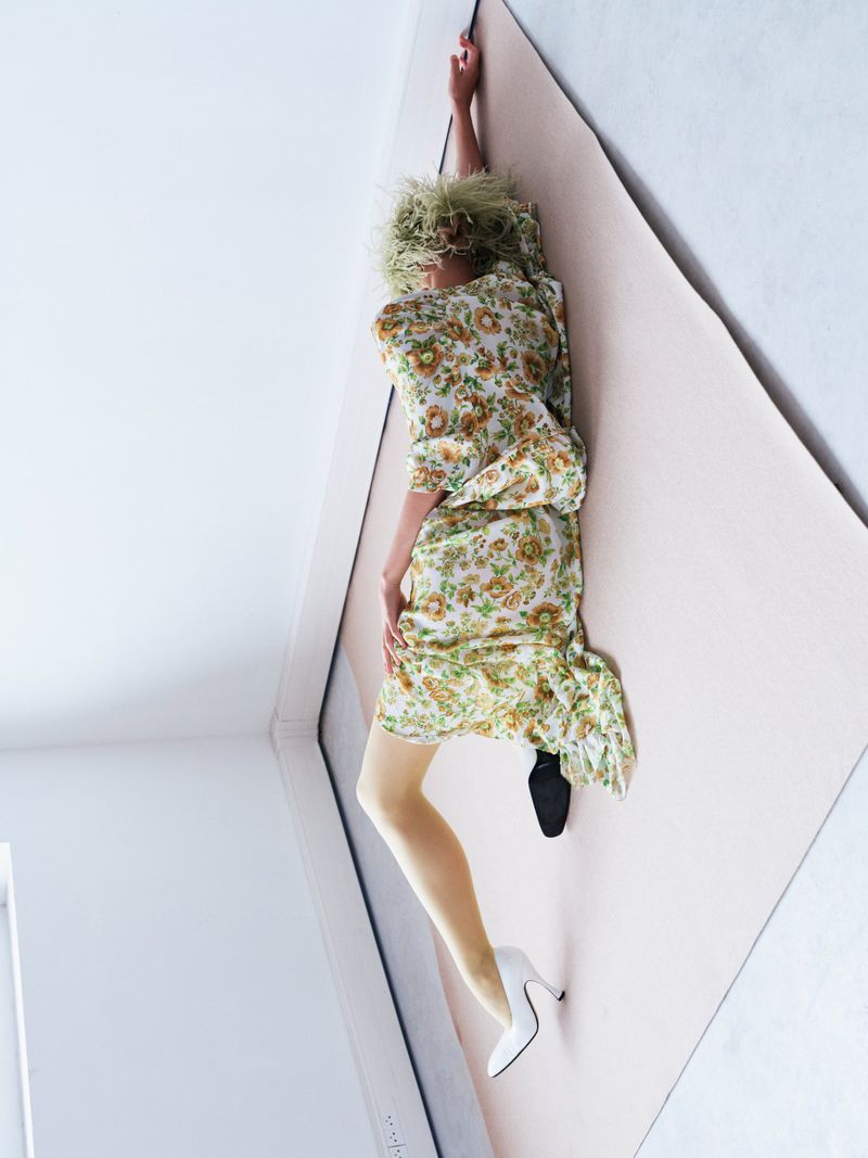 Woman in a floral dress, yellow tights, and mismatched shoes, one white and one black, lies on a light pink surface against a white wall.