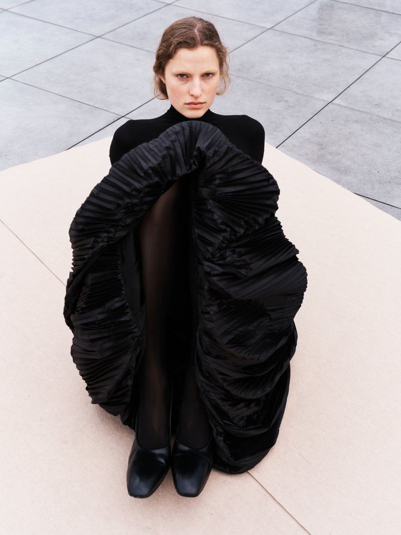 A woman in a black turtleneck and pleated skirt sits on a tiled surface, gazing directly at the camera with a serious expression.
