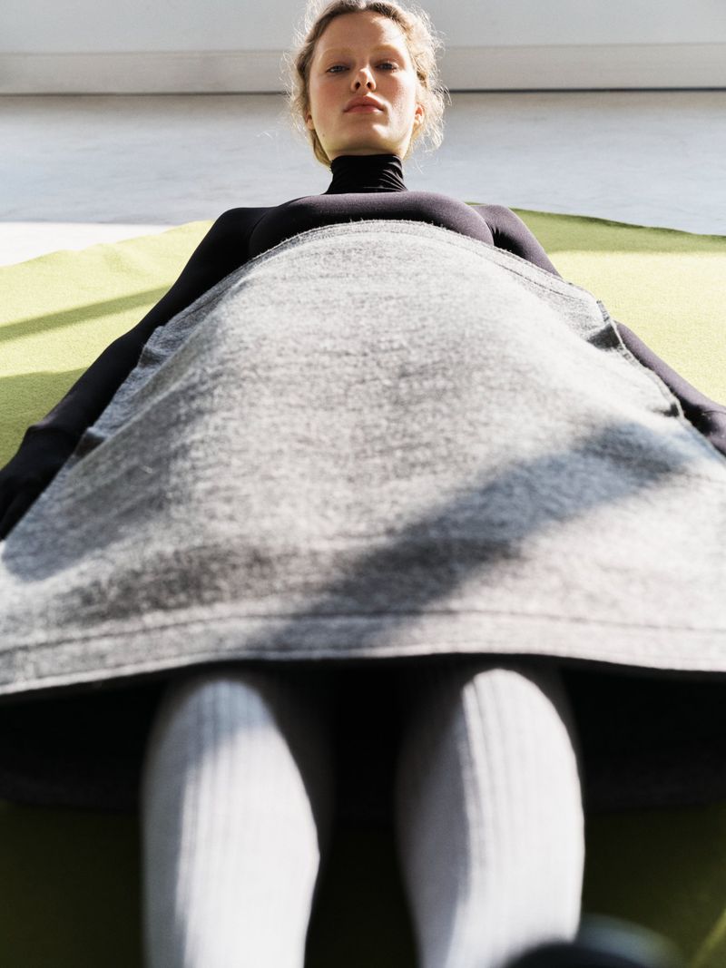 Eye-level view of a fair-skinned young woman in black turtleneck and gray dress, posing against a light green backdrop.
