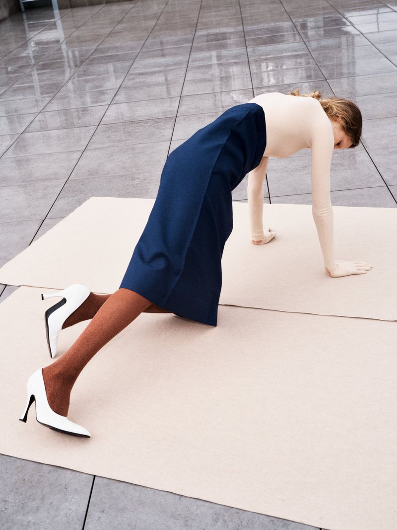 A woman in white top, navy skirt, and white high-heels does a plank on a cream-colored material surface in a bright indoor space.