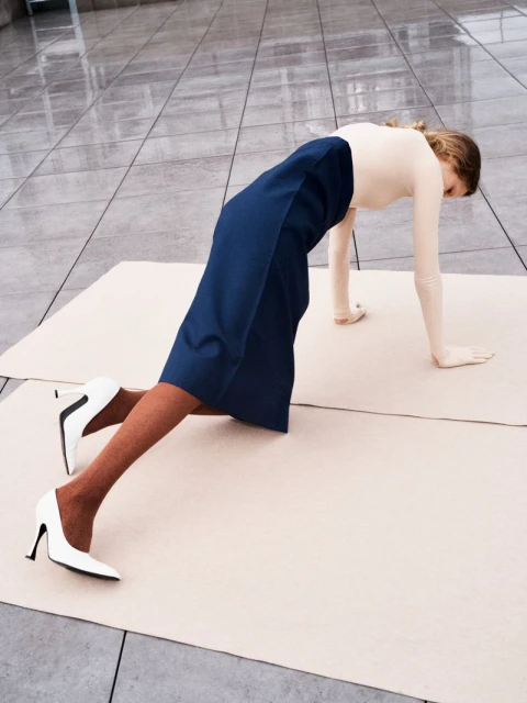 A woman in white top, navy skirt, and white high-heels does a plank on a cream-colored material surface in a bright indoor space.