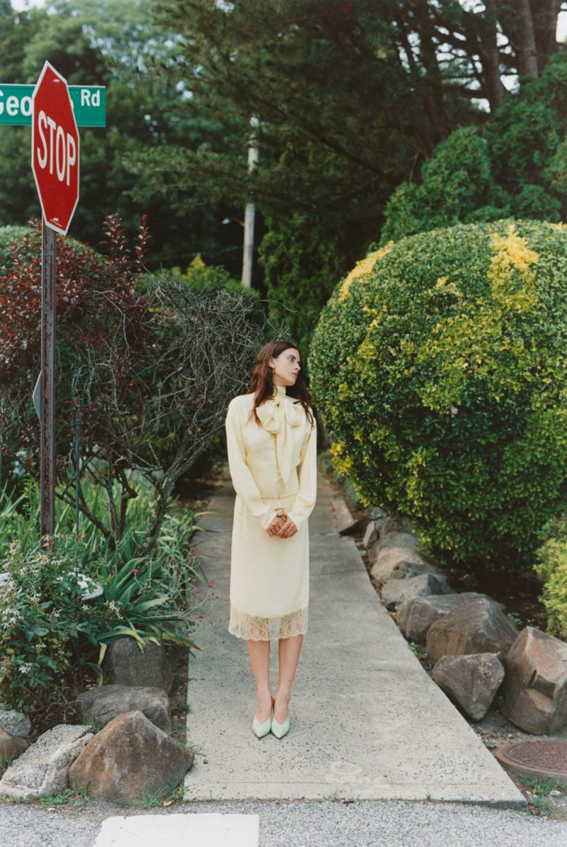 Woman in yellow dress poses near STOP sign in lush garden. Vintage fashion, outdoor setting with green tones, and natural lighting.