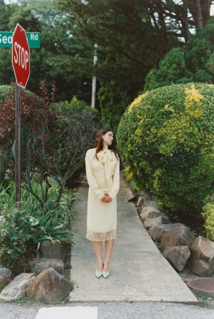 Woman in yellow dress poses near STOP sign in lush garden. Vintage fashion, outdoor setting with green tones, and natural lighting.
